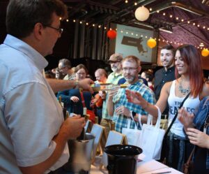 A man pouring wine for a woman at a crowded cheese and wine event